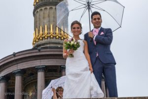 Hochzeitsfotograf Berlin - Junges Brautpaar unter einem durchsichtigen Regenschirm vor der Siegessäule in Berlin.